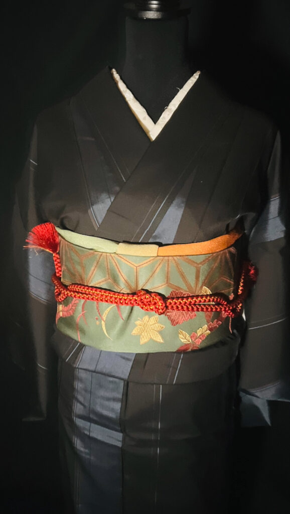黒の大島紬（単衣）の上半身。緑系の柄帯に赤の帯締めを合わせた帯まわりアップ（家紋なし）