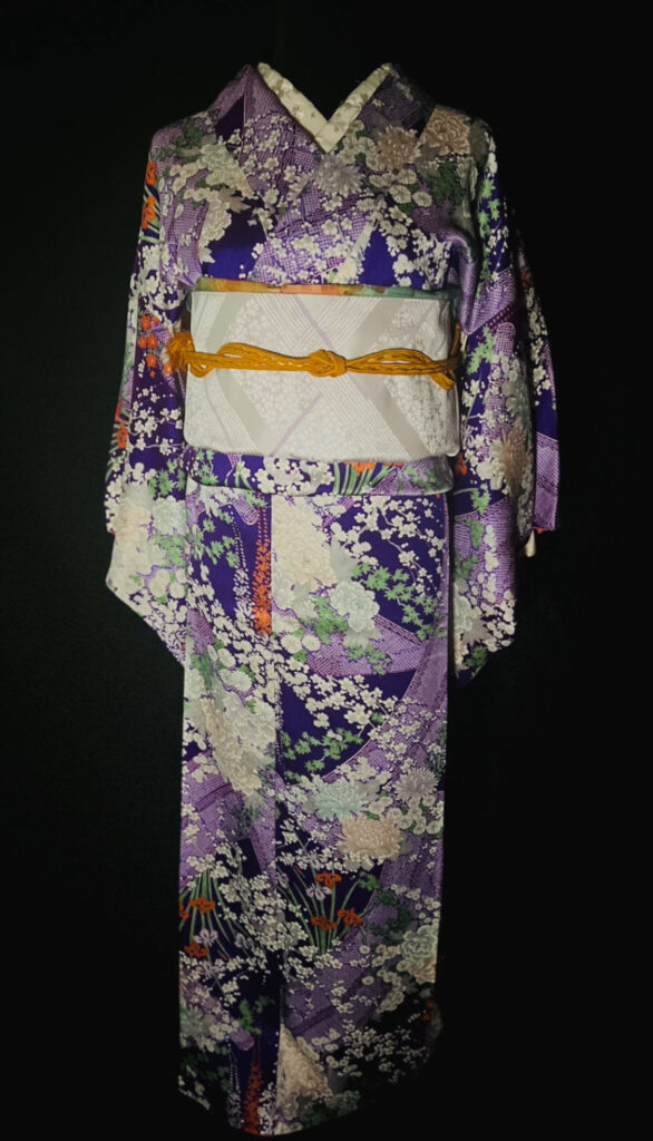 紫地に草花文様が広がる袷の正絹小紋。淡い名古屋帯とからし色の帯締めを合わせた全身コーディネート(家紋なし)