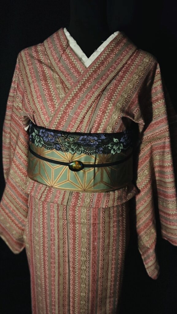 赤茶の縞模様ウール着物の胸元と帯まわり。緑×金の幾何学柄帯に、黒地の花柄帯揚げを合わせた着姿

