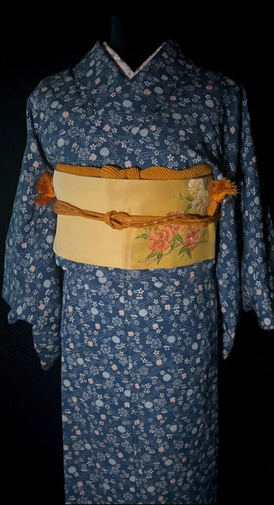 青の小花柄の小紋に、生成りの名古屋帯と黄土色の帯締めを合わせた全身写真