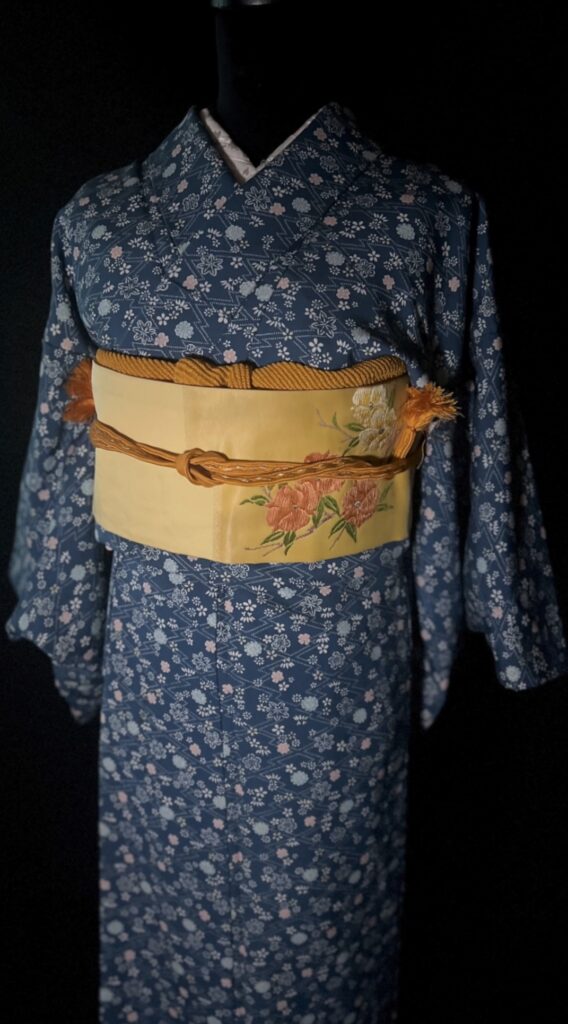 青の小紋の柄と、生成りの名古屋帯（花意匠）の組み合わせが分かる上半身写真